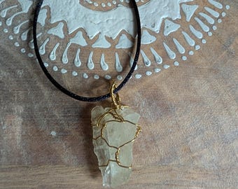 May include: A clear quartz crystal pendant wrapped in gold-colored wire, suspended from a black cord necklace. The pendant is rectangular and has a rough, natural texture. The necklace is displayed on a light-colored, patterned surface.
