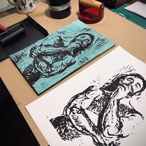 May include: A black and white linocut print of a person with their head resting on their hand. The print is on a white sheet of paper. The print is on a table with a blue and green linocut block in the background.