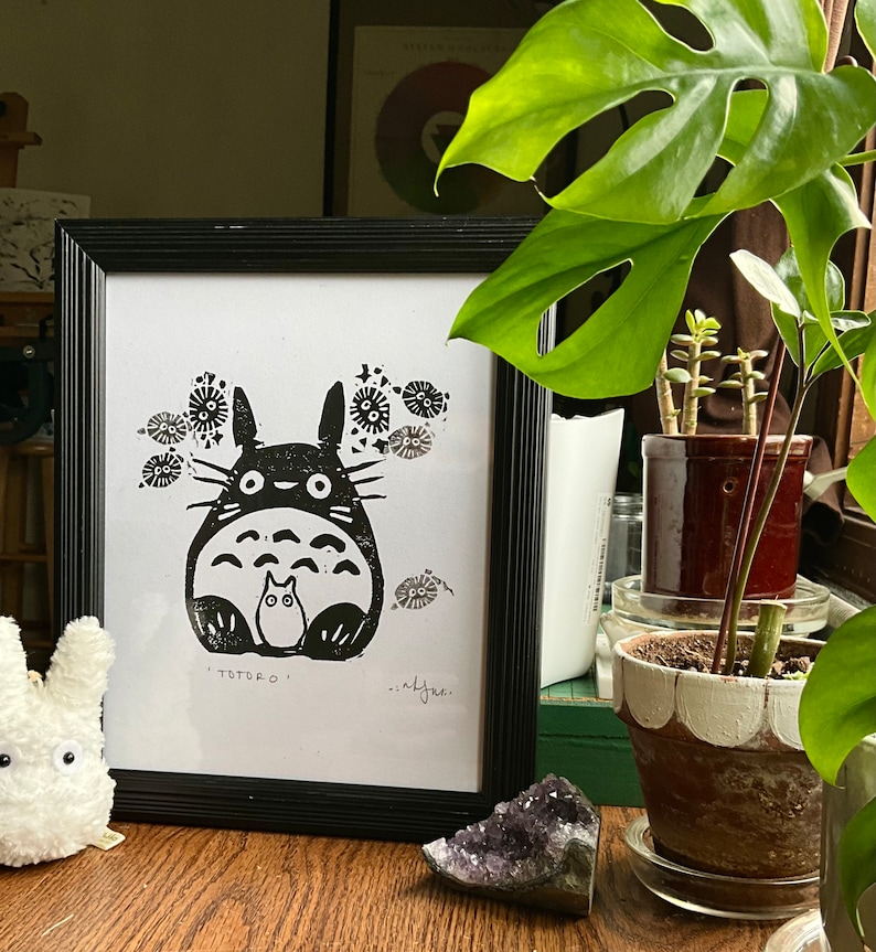 以下が含まれることがあります： 黒いフレームに入ったトトロのイラスト。このアートワークは、大きな体と2つの小さな姿を持つキャラクターを描いています。印刷は白い紙に印刷されており、画像の下に「TOTORO」という文字があります。印刷は木製の表面に表示されています。