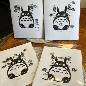 以下が含まれることがあります： 白黒のトトロのリノカットイラストの4つのプリント。各プリントには、小さなキャラクターと花のアクセントが付いたトトロのキャラクターが描かれています。「TOTORO」の文字が各プリントの下部に印刷されています。