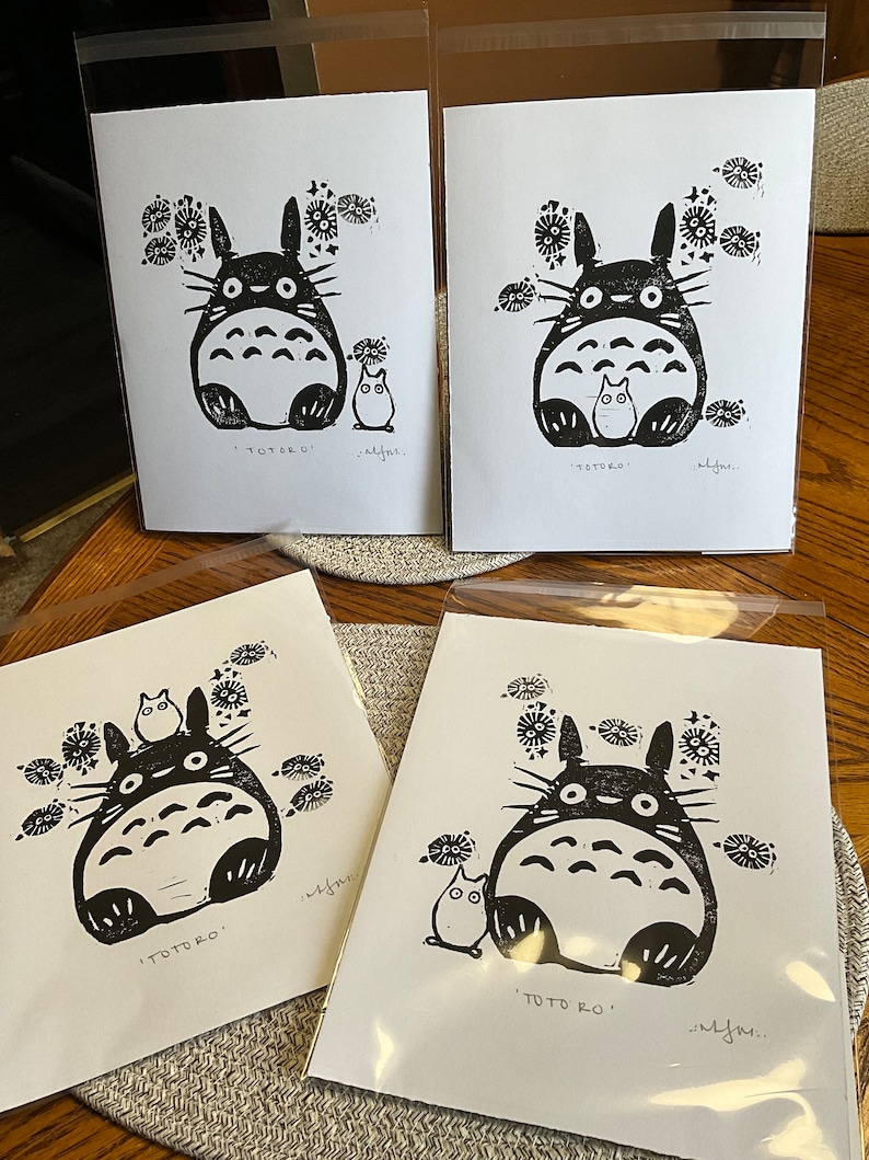 以下が含まれることがあります： 白黒のトトロのリノカットイラストの4つのプリント。各プリントには、小さなキャラクターと花のアクセントが付いたトトロのキャラクターが描かれています。「TOTORO」の文字が各プリントの下部に印刷されています。