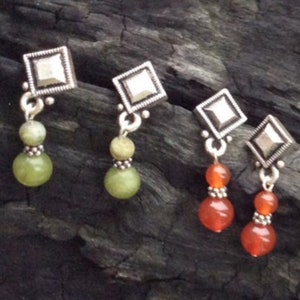 Puede incluir: Tres pares de pendientes con tapas cuadradas de plata y cuentas de piedras preciosas verdes y naranjas colgantes.