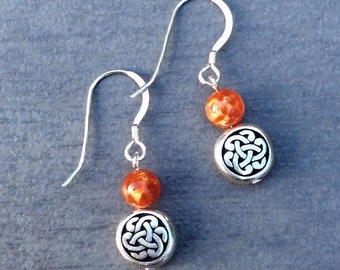May include: A pair of dangle earrings featuring silver-toned metal hooks, each suspending a single round orange marbled bead above a circular silver-toned charm embossed with an intricate, symmetrical Celtic-style knot pattern.