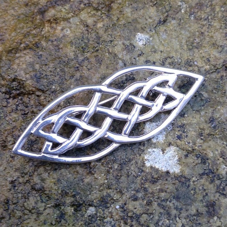 Pode incluir: Um broche de n&oacute; celta de prata com um design detalhado e intrincado. O broche tem a forma de uma folha e um acabamento liso e polido.