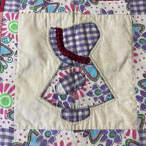 May include: A handmade quilt with a central appliqué of a dress shape in purple gingham and floral fabric. The quilt features a variety of patterned fabrics in shades of purple, pink, yellow, and white, with a scalloped edge.