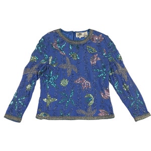Puede incluir: Top azul de manga larga con cuentas y cuello redondo. El top está adornado con lentejuelas en tonos azules, verdes, plateados y rosas, dispuestas en un patrón floral. El cuello y los puños están ribeteados con cuentas plateadas.