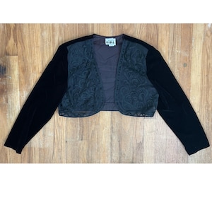 May include: A black velvet bolero jacket with long sleeves. The jacket features a dark gray embroidered floral design on the front and the brand name "niki" on a tag.
