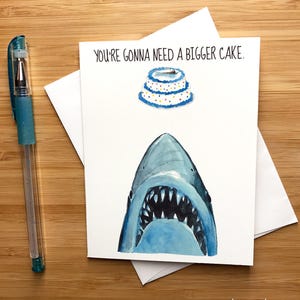 May include: A birthday card with a shark's mouth open wide and a cake above it. The card reads "You're gonna need a bigger cake."