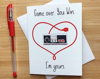 May include: A white card with red text that reads "Game over. You Win." and a red heart shape with a red cord and a black and red video game controller inside. The text "I'm yours." is below the heart. The card has a website address at the bottom.