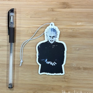 May include: A white car air freshener with a black and white image of a character with a spiked head and a black outfit. The character is holding their hands out in front of them. The text "http://yeaohgreetings.com" is printed at the bottom of the air freshener.