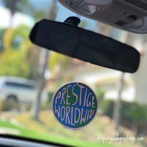 May include: A round car air freshener with a blue and green globe design. The text "PRESTIGE WORLDWIDE" is written in white on the globe. The air freshener is hanging from the rearview mirror of a car.