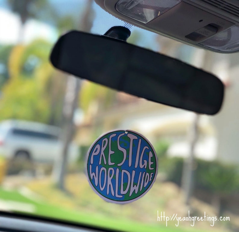 May include: A round car air freshener with a blue and green globe design. The text "PRESTIGE WORLDWIDE" is written in white on the globe. The air freshener is hanging from the rearview mirror of a car.
