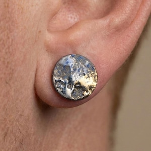 Könnte beinhalten: Ein silberfarbener, strukturierter Ohrstecker mit kreisförmigem Design. Der Ohrring hat ein gehämmertes Metallaussehen mit Andeutungen von Blau und Gold. Der Ohrring wird in einem Ohrloch getragen.