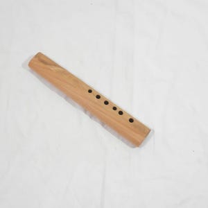 Könnte beinhalten: Eine hellbraune Holzflöte mit sieben Fingerlöchern. Das Instrument ist rechteckig mit abgerundeten Kanten und einer glatten Oberfläche. Die Flöte liegt auf einer weißen Oberfläche.