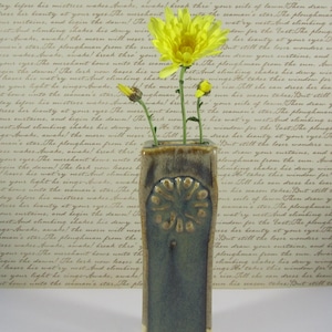 May include: A rectangular ceramic vase with a floral design, holding yellow flowers. The vase has a textured, mottled green and brown glaze. The background features faded script.