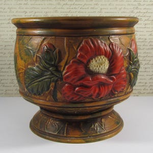 May include: A decorative, brown ceramic planter with a pedestal base. The planter features a raised relief design of a red poppy flower with green leaves. The planter is approximately 7 inches tall and 7 inches wide.