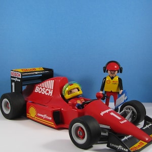 May include: A red toy race car with black tires and sponsor logos, including "Bosch" and "Goodyear." A driver figure in a helmet sits in the cockpit. A second figure in a yellow vest and headset stands nearby. The car has the number "1."