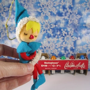May include: A blue and red felt Christmas ornament elf with a white fur trim and closed eyes. The elf is holding its hands together in front of its chest. The ornament is hanging from a gold string. The elf is in front of a box of Westinghouse Christmas bulbs.