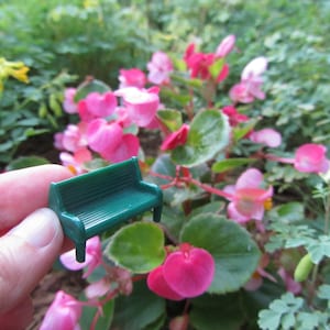 May include: A miniature green plastic bench sits on a hand in front of a bed of pink flowers. The flowers are in bloom and have green leaves.