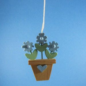 May include: A wooden flower pot ornament with three gray gear-shaped flowers, green leaves, and a blue heart. The pot is brown and suspended by a beige string against a blue background. A charming, handcrafted decoration.