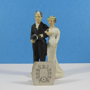 May include: A vintage wedding cake topper featuring a bride and groom figurine. The groom wears a black tuxedo and holds a hat. The bride is in a white dress. A watch face with numbers is in the foreground. The background is a solid blue.