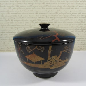 May include: A black, lidded bowl with a pedestal base. The bowl features a gold and red landscape design with a pagoda, trees, and floral accents. The lid has a small black knob and a blue rim.