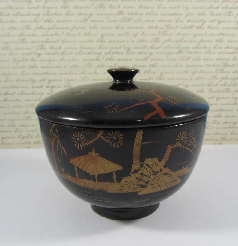 May include: A black, lidded bowl with a pedestal base. The bowl features a gold and red landscape design with a pagoda, trees, and floral accents. The lid has a small black knob and a blue rim.