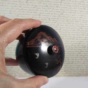 May include: A black, oval container with a decorative lid. The lid features a mountain scene in shades of brown and red, with a small, round knob adorned with a floral design. The container is held in a hand.