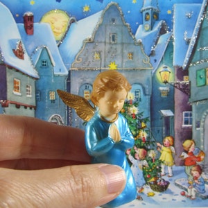 May include: A blue angel figurine with gold wings, holding its hands together in prayer. The figurine is in front of a vintage Christmas advent calendar with a snowy village scene.