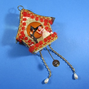 May include: A small, red and gold cuckoo clock ornament with a white face and a couple inside. The clock has a pendulum with a gold chain and two white beads.