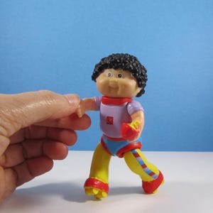 May include: A colorful Cabbage Patch Kids figurine. The doll has black curly hair, a light skin tone, and is wearing a purple shirt, red collar, blue shorts, yellow pants, and red shoes. The doll is in a running pose.