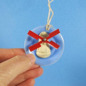 May include: A blue and white plastic ornament with a red windmill design. The ornament is shaped like a circle and has a string attached to the top.
