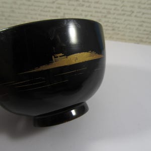 May include: A black bowl with a rounded shape and a small, circular base. The bowl is decorated with a gold-coloured, abstract design. It has a glossy finish and appears to be made of a hard material.