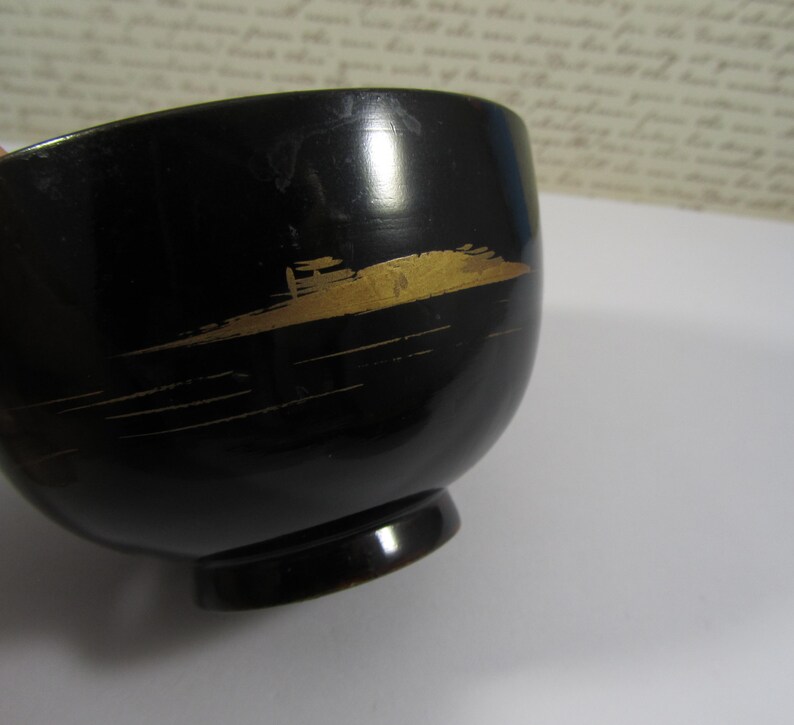 May include: A black bowl with a rounded shape and a small, circular base. The bowl is decorated with a gold-coloured, abstract design. It has a glossy finish and appears to be made of a hard material.