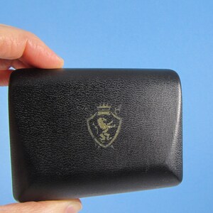May include: A black, textured box with a gold emblem featuring a lion and crown design. The box is held in a hand against a blue background. The box appears to be made of leather or a similar material.