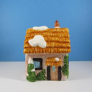 May include: A ceramic cottage figurine featuring a textured orange thatched roof with white snow accents, a light pink facade, a brown door, a small window, and green foliage. The structure includes a chimney and a small snow-covered porch.