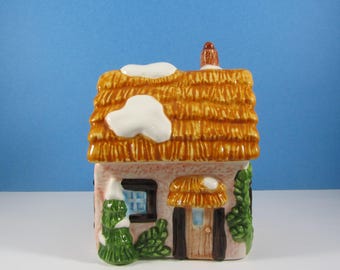 May include: A ceramic cottage figurine featuring a textured orange thatched roof with white snow accents, a light pink facade, a brown door, a small window, and green foliage. The structure includes a chimney and a small snow-covered porch.