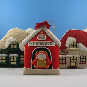 May include: Three felt Christmas ornaments shaped like houses. The center ornament is red and white and says "Firehouse No. 3" on the front. The ornament on the left is green and white and says "Baker" on the front. The ornament on the right is white and red and says "Barber" on the front.
