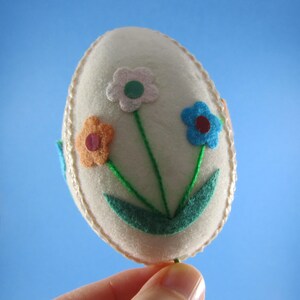 May include: A cream-colored felt Easter egg decoration with three colorful felt flowers. The flowers are white, orange, and blue, with green stems and leaves. The egg is trimmed with a gold-colored thread.