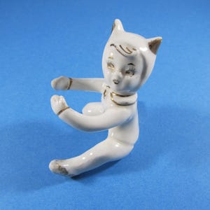 May include: A white ceramic figurine of a seated cat-like figure with gold accents on the ears, collar, and arms. The figurine has closed eyes and outstretched arms, set against a blue background.