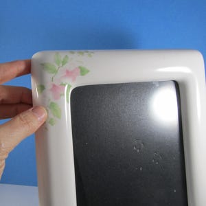 May include: White rectangular picture frame with a floral design in the upper left corner. The flowers are pink and the leaves are green. The frame is held by a hand.