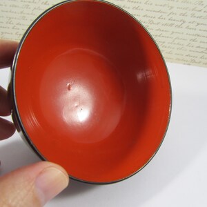 May include: A small, round bowl with a bright red interior and a dark, possibly black, exterior rim. The bowl's interior is glossy, reflecting light, and the rim has a thin, metallic edge. The bowl is held by a hand.