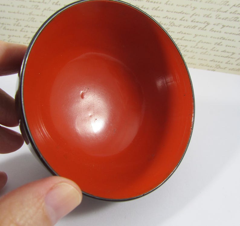 May include: A small, round bowl with a bright red interior and a dark, possibly black, exterior rim. The bowl's interior is glossy, reflecting light, and the rim has a thin, metallic edge. The bowl is held by a hand.