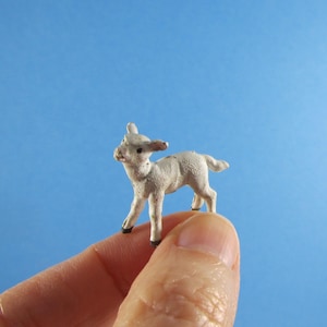 May include: A small, off-white lamb figurine with black hooves stands on a finger against a blue background. The lamb has detailed features, including ears and a tail. This miniature lamb is a charming decorative item.
