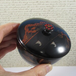 May include: A black, oval trinket box with a lid. The box has orange and white decorative elements, including a floral design on the lid's knob. The box is held in a hand.