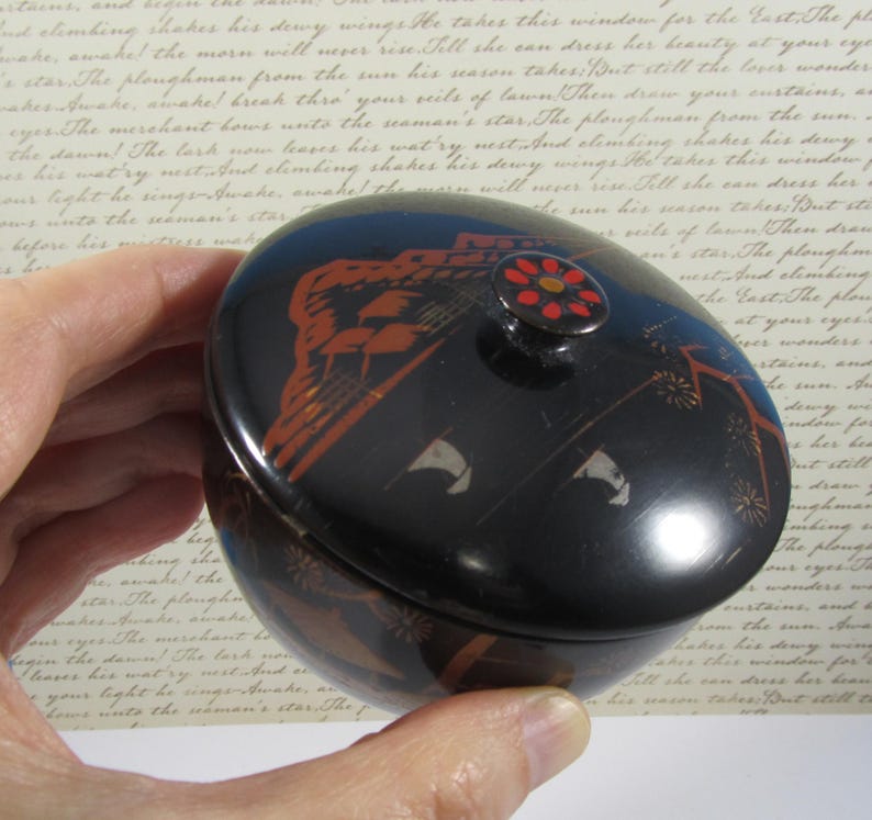May include: A black, oval trinket box with a lid. The box has orange and white decorative elements, including a floral design on the lid's knob. The box is held in a hand.