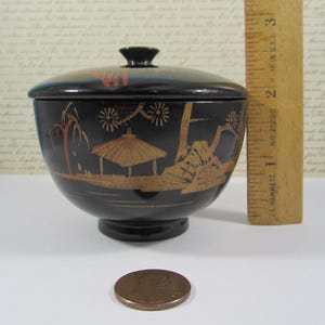 May include: A black, lidded bowl with a pedestal base, featuring a gold and red landscape design. The bowl is approximately 5 cm in diameter. A penny is next to the bowl for size comparison.
