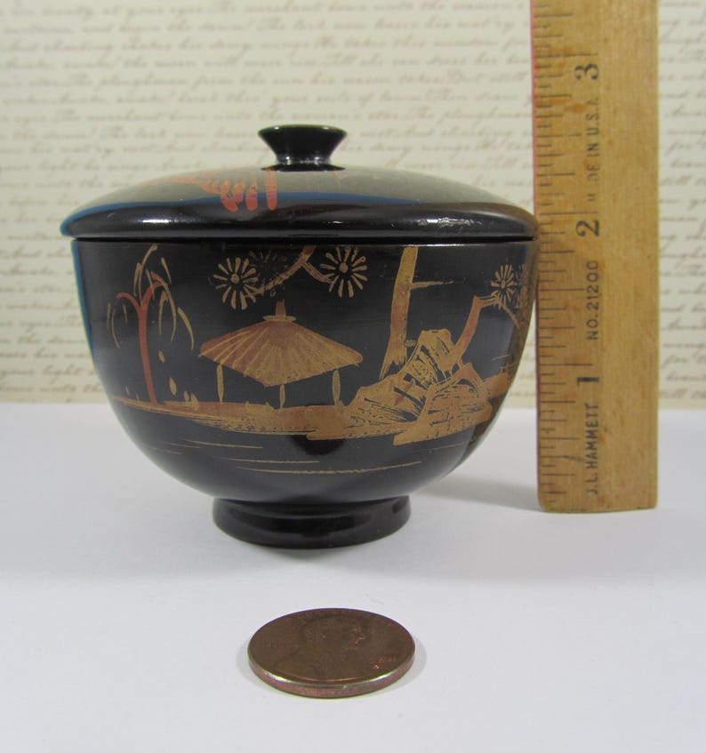 May include: A black, lidded bowl with a pedestal base, featuring a gold and red landscape design. The bowl is approximately 5 cm in diameter. A penny is next to the bowl for size comparison.