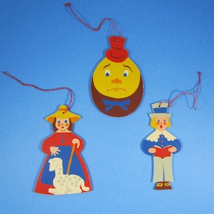 May include: Three wooden ornaments with a red string for hanging. The first ornament is a yellow face with a blue bow tie and a red hat. The second ornament is a girl with a red dress, a yellow hat, and a white lamb. The third ornament is a boy with a blue and white outfit, a red book, and a white hat.