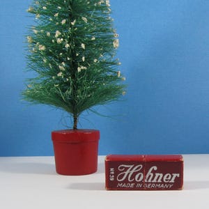 May include: A small, artificial Christmas tree in a red pot stands next to a red rectangular box. The box has the word "Hohner" in white script, with "MADE IN GERMANY" below. The tree is green with white accents.